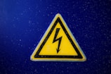 A triangular yellow warning sign with a black lightning bolt symbol in the center is mounted on a blue background. The sign is designed to indicate electrical hazard or high voltage.