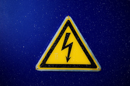 A triangular yellow warning sign with a black lightning bolt symbol in the center is mounted on a blue background. The sign is designed to indicate electrical hazard or high voltage.