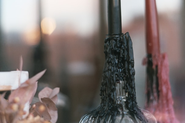 Melted wax drips down the sides of two candlesticks placed in glass holders. One candle is black while the other is red, creating an interesting contrast. In the foreground, dried flowers add a rustic and vintage touch to the scene. The background is softly blurred, emphasizing the candles and flowers.