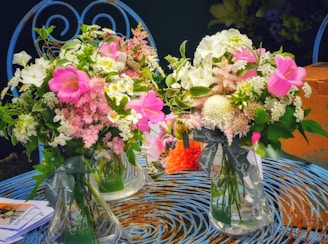Two glass vases filled with lush bouquets of flowers featuring bright pink, white, and green hues sit on an intricately designed blue metal table. The flowers include varieties like roses, daisies, and carnations with abundant green foliage. Complementing this scene is a soft orange flower in the background, and an adjacent brown cardboard box, as well as some printed paper with visible text.