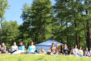 A group of people is sitting cross-legged on grass in a forested area, engaged in meditation. In the background, there is a traditional yurt among tall green trees. The sky is clear and blue, suggesting a sunny day.