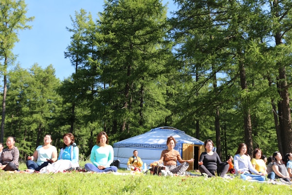 A group of people is sitting cross-legged on grass in a forested area, engaged in meditation. In the background, there is a traditional yurt among tall green trees. The sky is clear and blue, suggesting a sunny day.
