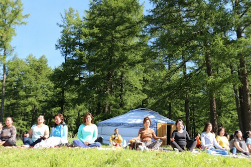 A group of people is sitting cross-legged on grass in a forested area, engaged in meditation. In the background, there is a traditional yurt among tall green trees. The sky is clear and blue, suggesting a sunny day.