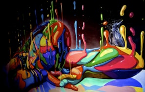 A vibrant, colorful meld of dripping paint forms the shape of a reclining human figure, with a small bird perched on its side. The combination of fluid, bright paint creates an abstract and dynamic composition against a dark background.