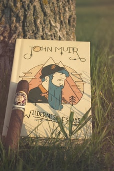 A book titled 'John Muir Wilderness Essays' is propped against the trunk of a tree. The cover features an illustration of a stylized man with mountains in the background. A cigar with a decorative label is placed next to the book. Grass surrounds the scene, suggesting an outdoor setting.