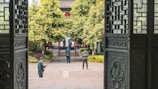 A serene courtyard scene framed by intricately designed wooden doors. Two individuals are standing; one person appears to be a guard in uniform, and another casually dressed person is using a phone. Lush green trees and traditional architectural elements enhance the background.
