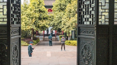 A serene courtyard scene framed by intricately designed wooden doors. Two individuals are standing; one person appears to be a guard in uniform, and another casually dressed person is using a phone. Lush green trees and traditional architectural elements enhance the background.