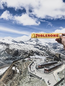 A person holds a Toblerone chocolate bar in the foreground with a scenic view of snow-capped mountains and partly cloudy skies in the background. Below, tourists are gathered on a large viewing platform near a train station, featuring orange trains.