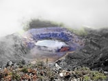 A volcanic crater filled with blue and green sulfuric minerals surrounds a lake of water. The landscape is covered in mist and clouds, with rugged terrain and exposed rock formations. Vegetation borders the bottom edge, contrasting against the crater's stark colors.