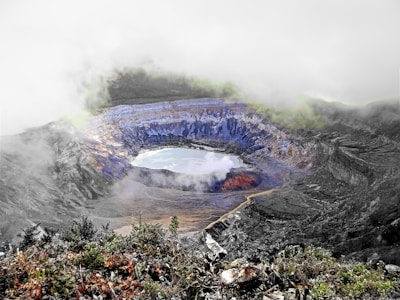 A volcanic crater filled with blue and green sulfuric minerals surrounds a lake of water. The landscape is covered in mist and clouds, with rugged terrain and exposed rock formations. Vegetation borders the bottom edge, contrasting against the crater's stark colors.