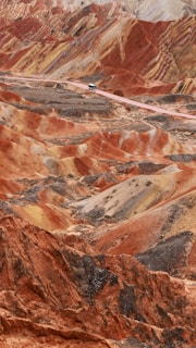 A vibrant and colorful landscape features layered hills and mountains with distinct striations of red, orange, and yellow hues. The terrain is rugged and undulating with a lone bus traveling on a narrow road that winds through the unique geologic formations.