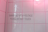 A tiled wall with a warning sign stating '!WARNING! HIGH VOLTAGE PLEASE DO NOT TOUCH'. The tiles have a glossy finish and a soft pink hue due to lighting. There is a round light bulb partially visible in the top right corner.