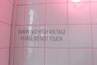 A tiled wall with a warning sign stating '!WARNING! HIGH VOLTAGE PLEASE DO NOT TOUCH'. The tiles have a glossy finish and a soft pink hue due to lighting. There is a round light bulb partially visible in the top right corner.