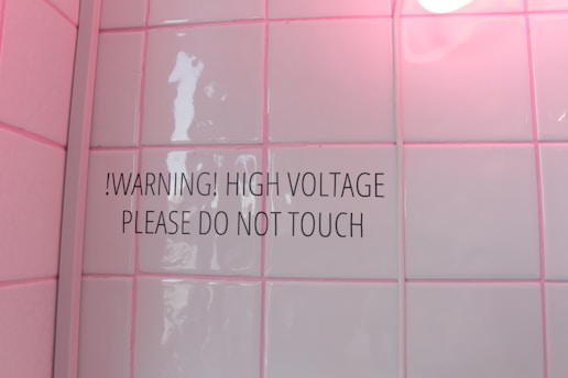 A tiled wall with a warning sign stating '!WARNING! HIGH VOLTAGE PLEASE DO NOT TOUCH'. The tiles have a glossy finish and a soft pink hue due to lighting. There is a round light bulb partially visible in the top right corner.