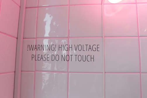 A tiled wall with a warning sign stating '!WARNING! HIGH VOLTAGE PLEASE DO NOT TOUCH'. The tiles have a glossy finish and a soft pink hue due to lighting. There is a round light bulb partially visible in the top right corner.