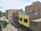 A residential urban neighborhood with multi-story buildings. The structures are mainly constructed with brick and concrete, featuring colorful facades including yellow and blue. Some buildings have visible unfinished brickwork. Trees and plants line the street below, and laundry hangs from a rooftop. There are a few cars parked and a couple of people walking along the street.