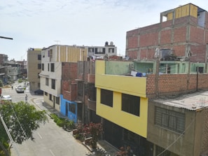 A residential urban neighborhood with multi-story buildings. The structures are mainly constructed with brick and concrete, featuring colorful facades including yellow and blue. Some buildings have visible unfinished brickwork. Trees and plants line the street below, and laundry hangs from a rooftop. There are a few cars parked and a couple of people walking along the street.