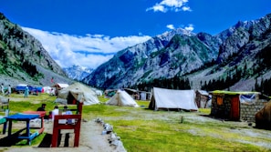 A picturesque mountain landscape featuring several tents set up on a grassy plain. The scene includes snow-capped mountains in the background and a bright blue sky with scattered clouds. People are sitting around tables, and the area is surrounded by lush green forests.