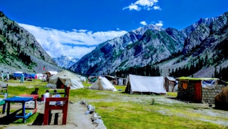 A picturesque mountain landscape featuring several tents set up on a grassy plain. The scene includes snow-capped mountains in the background and a bright blue sky with scattered clouds. People are sitting around tables, and the area is surrounded by lush green forests.