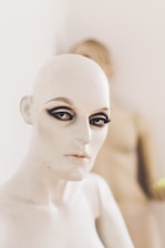 A mannequin with a bald head and prominent makeup, featuring dark eyeshadow and eyeliner, is positioned in soft focus. Another mannequin appears blurred in the background, adding depth to the scene. The lighting is soft and neutral.