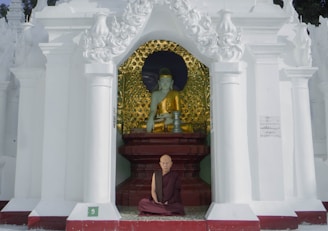 A monk sits cross-legged in front of a statue in a temple alcove, adorned with ornate white pillars and intricate designs. The statue is of a serene figure dressed in golden robes against a background with golden patterns.