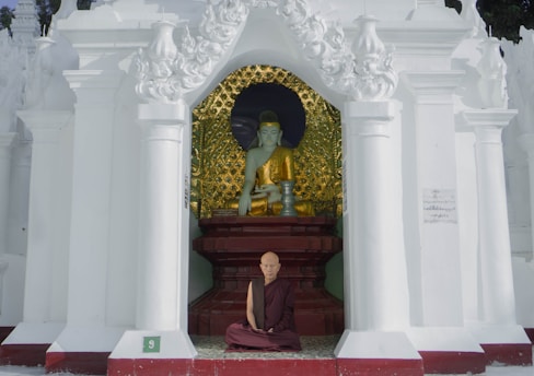 A monk sits cross-legged in front of a statue in a temple alcove, adorned with ornate white pillars and intricate designs. The statue is of a serene figure dressed in golden robes against a background with golden patterns.