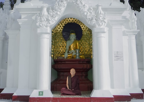 A monk sits cross-legged in front of a statue in a temple alcove, adorned with ornate white pillars and intricate designs. The statue is of a serene figure dressed in golden robes against a background with golden patterns.
