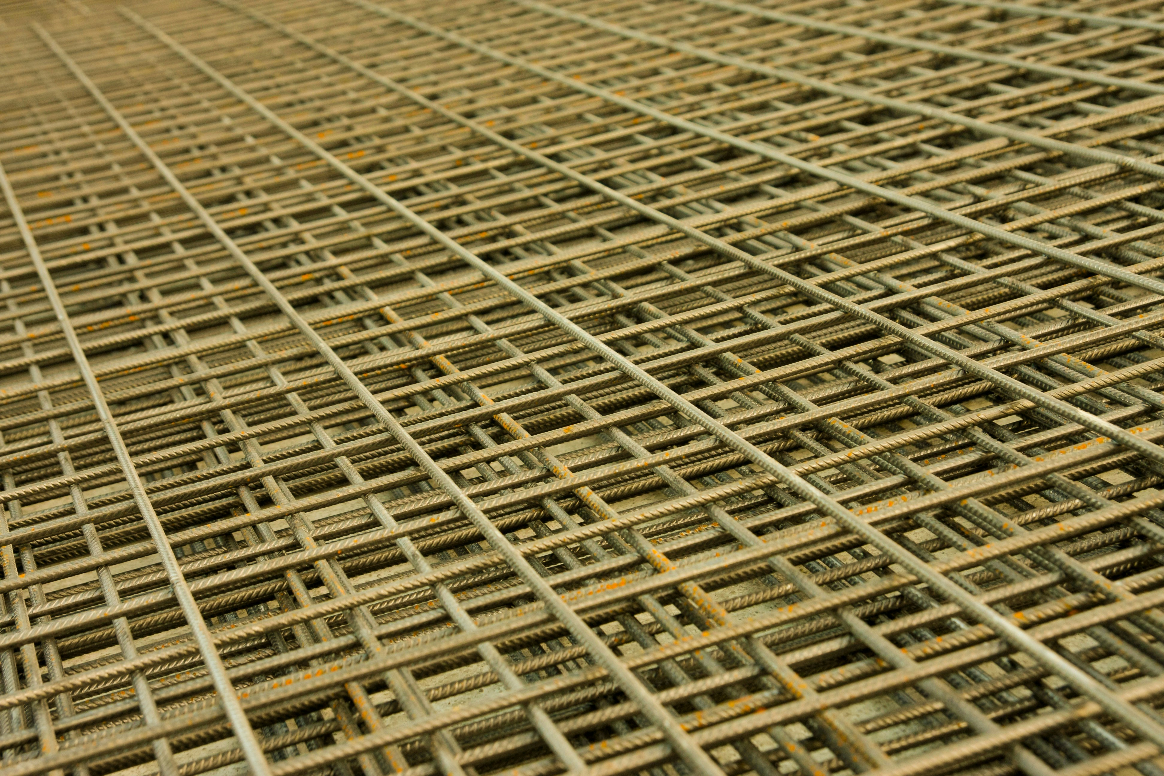 An intricate pattern of intersecting steel rebar forming a mesh-like structure. The grid is composed of parallel and perpendicular bars creating a complex, orderly framework typically used in construction for reinforcement.