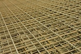 An intricate pattern of intersecting steel rebar forming a mesh-like structure. The grid is composed of parallel and perpendicular bars creating a complex, orderly framework typically used in construction for reinforcement.