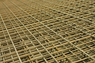 An intricate pattern of intersecting steel rebar forming a mesh-like structure. The grid is composed of parallel and perpendicular bars creating a complex, orderly framework typically used in construction for reinforcement.
