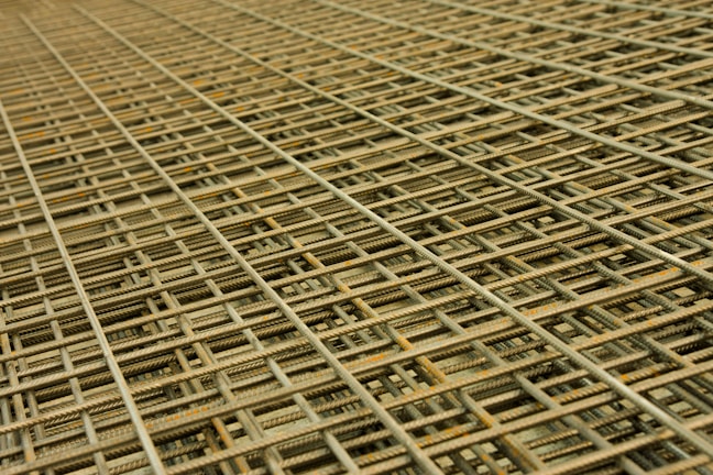 An intricate pattern of intersecting steel rebar forming a mesh-like structure. The grid is composed of parallel and perpendicular bars creating a complex, orderly framework typically used in construction for reinforcement.