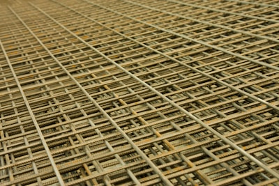 An intricate pattern of intersecting steel rebar forming a mesh-like structure. The grid is composed of parallel and perpendicular bars creating a complex, orderly framework typically used in construction for reinforcement.