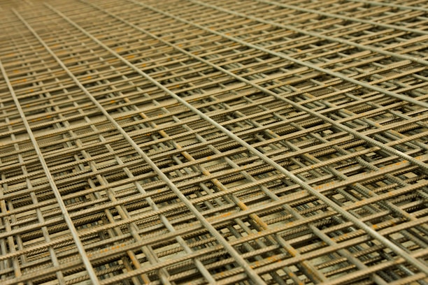 An intricate pattern of intersecting steel rebar forming a mesh-like structure. The grid is composed of parallel and perpendicular bars creating a complex, orderly framework typically used in construction for reinforcement.