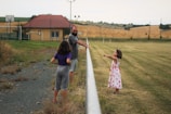 A man and two children are outdoors near a grassy field and a pathway. The children interact playfully, one wearing a floral dress and the other in a purple shirt. A house with a red roof is visible in the background, along with tilled fields and a few trees.