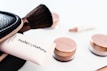 A collection of cosmetics is neatly displayed, including a makeup brush, a black zippered pouch, and several rose-gold capped jars. A tube that reads 'nude by nature' is partially visible, suggesting a beauty product brand.