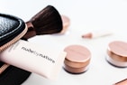 A collection of cosmetics is neatly displayed, including a makeup brush, a black zippered pouch, and several rose-gold capped jars. A tube that reads 'nude by nature' is partially visible, suggesting a beauty product brand.