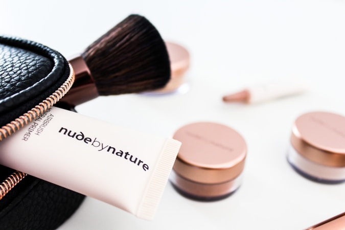 A collection of cosmetics is neatly displayed, including a makeup brush, a black zippered pouch, and several rose-gold capped jars. A tube that reads 'nude by nature' is partially visible, suggesting a beauty product brand.