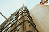 Close-up view of metal rebar protruding from a concrete structure, arranged in a grid pattern. The perspective is angled upward, giving a sense of height and construction progress. The sky in the background is clear, providing a contrast to the industrial materials.