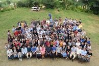 A group photo of diverse community members seated comfortably on the grass, framed by gentle greenery.
