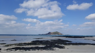A serene beach scene featuring a mix of sand and black lava rocks in the foreground. The ocean stretches out with a small, green hilly island in the distance. Above, the sky is bright with scattered fluffy clouds.