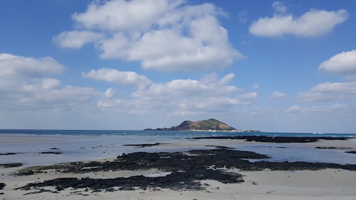 A serene beach scene featuring a mix of sand and black lava rocks in the foreground. The ocean stretches out with a small, green hilly island in the distance. Above, the sky is bright with scattered fluffy clouds.