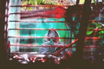 Graffiti art featuring a hooded figure with hands in a prayer position, set against a background with a teal and green color palette. The image is partially obscured by wooden slats, and there are vivid red and orange hues present, possibly representing either lighting or painted elements. The setting appears to be outdoors with trees and foliage visible in the background.