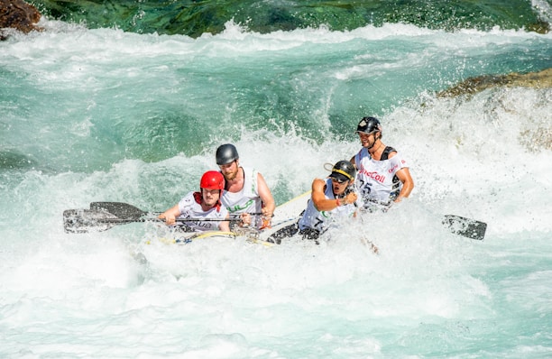 A group of four people are intensely focused as they navigate a raft through powerful whitewater rapids. They are wearing protective helmets and life jackets, showing determination and teamwork as they paddle through the churning water.