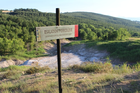 A warning sign is prominently displayed in the foreground, reading 'ESALAZIONI PERICOLOSE' which translates to 'Dangerous Fumes'. This is set against a backdrop of lush green forested hills and a clear sky, with some dry stream or riverbeds meandering through the landscape.