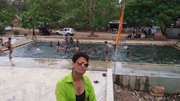 A group of people enjoying a swim in a rectangular outdoor pool surrounded by trees and greenery. In the foreground, a person wearing sunglasses and a bright green shirt is sitting on a ledge, and an orange flag is visible near the pool. In the background, there are parked cars and motorbikes, along with more people walking around.
