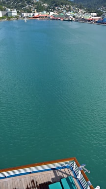 A serene body of water stretches out with a vibrant teal hue, bordered by a distant shoreline dotted with colorful buildings and lush greenery. In the foreground, part of a ship's deck with green deck chairs is visible, hinting at a relaxing cruise experience.