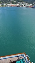 A serene body of water stretches out with a vibrant teal hue, bordered by a distant shoreline dotted with colorful buildings and lush greenery. In the foreground, part of a ship's deck with green deck chairs is visible, hinting at a relaxing cruise experience.