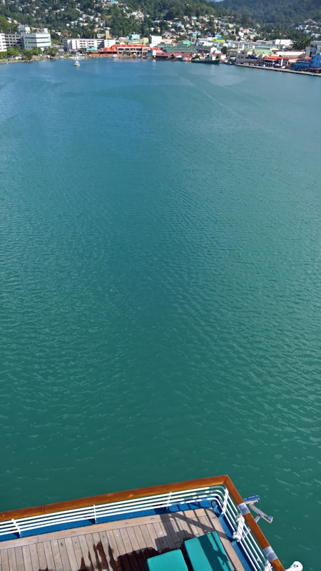 A serene body of water stretches out with a vibrant teal hue, bordered by a distant shoreline dotted with colorful buildings and lush greenery. In the foreground, part of a ship's deck with green deck chairs is visible, hinting at a relaxing cruise experience.