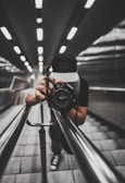 A person wearing a cap is holding a camera, capturing a shot while leaning down on a modern, sleek staircase with metallic railings. The perspective creates a dynamic, symmetrical view along the lines of the railings and the tiles, with ceiling lights adding depth.