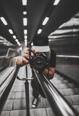 A person wearing a cap is holding a camera, capturing a shot while leaning down on a modern, sleek staircase with metallic railings. The perspective creates a dynamic, symmetrical view along the lines of the railings and the tiles, with ceiling lights adding depth.
